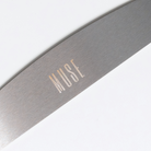 A metal reusable nail file with the branding 'MUSE' on it, lying on a white surface.